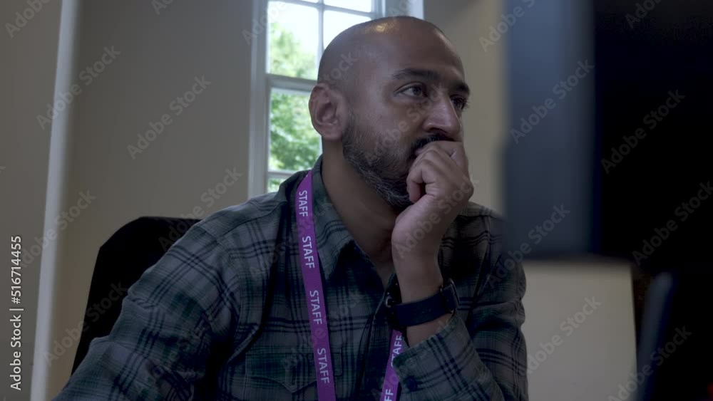 A close up shot from behind an office computer as an Asian Indian man sits with a clutched hand covering his mouth while reading the content displayed on a desktop monitor A close up shot from behind an office computer as an Asian Indian man sits with a clutched hand covering his mouth while reading the content displayed on a desktop monitor