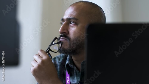 A close up shot from behind an office computer as an Asian man sits gazing into the air in deep thought about an issue while subconsciously tapping his spectacles on his chin photo