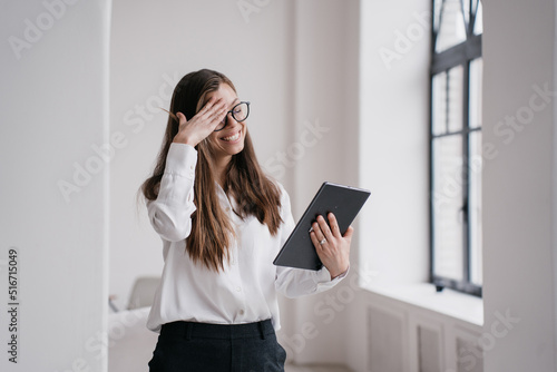 Grateful young businesswoman eyes closed touching her head in excited expression talking by tablet making video call, received great news. Satisfied girl in white shirt, black pants remote studying.
