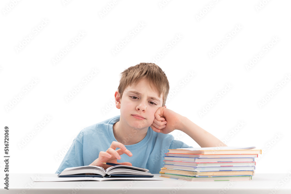 Tired boy doing homework at table and attentively reads book. Portrait of schoolboy at desk with stack of notebooks. White background. Tired boy doing homework at table and attentively reads book. Portrait of schoolboy at desk with stack of notebooks. White background.