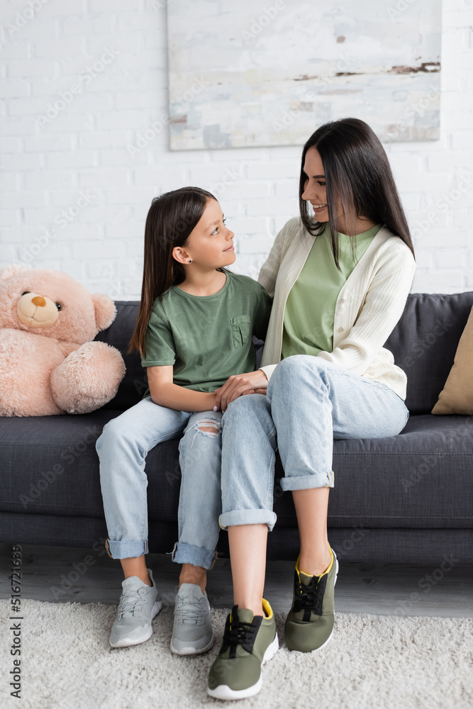 Φωτογραφία full length of girl and nanny sitting on couch near teddy bear and smiling at each other