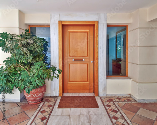 A contemporary house door made of natural wood and two windows, with a decorative potted plant. Athens, Greece.
