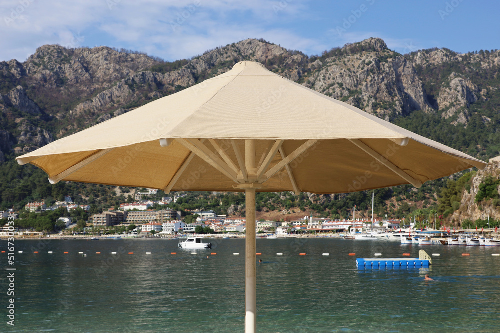 Beach vacation, sun umbrella on sea background. View to mountains on Mediterranean coast, tourist boats and resort town