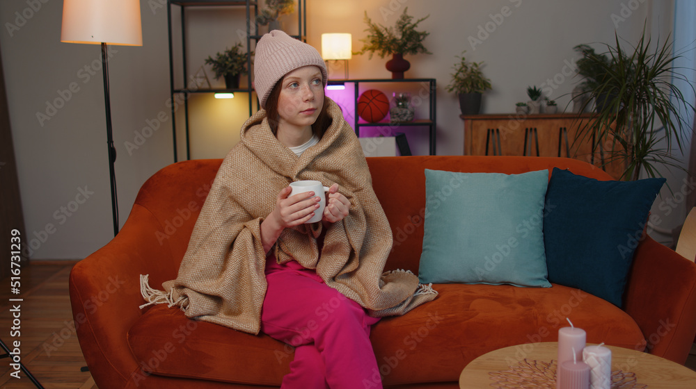 Obraz premium Sick child girl wear hat wrapped in plaid sit alone shivering from cold on sofa drinking hot tea in unheated apartment without heating due debt. Unhealthy kid feeling discomfort try to warming up