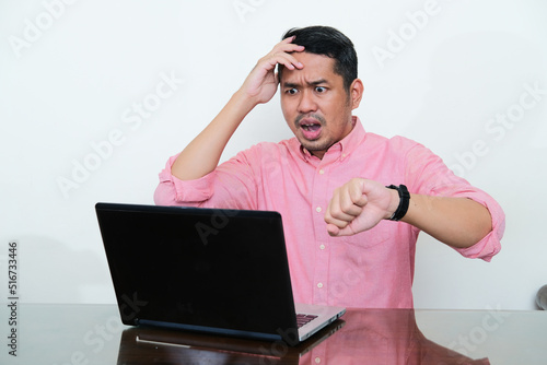 Adult Asian man looking to his laptop with worried expression while showing his watch photo