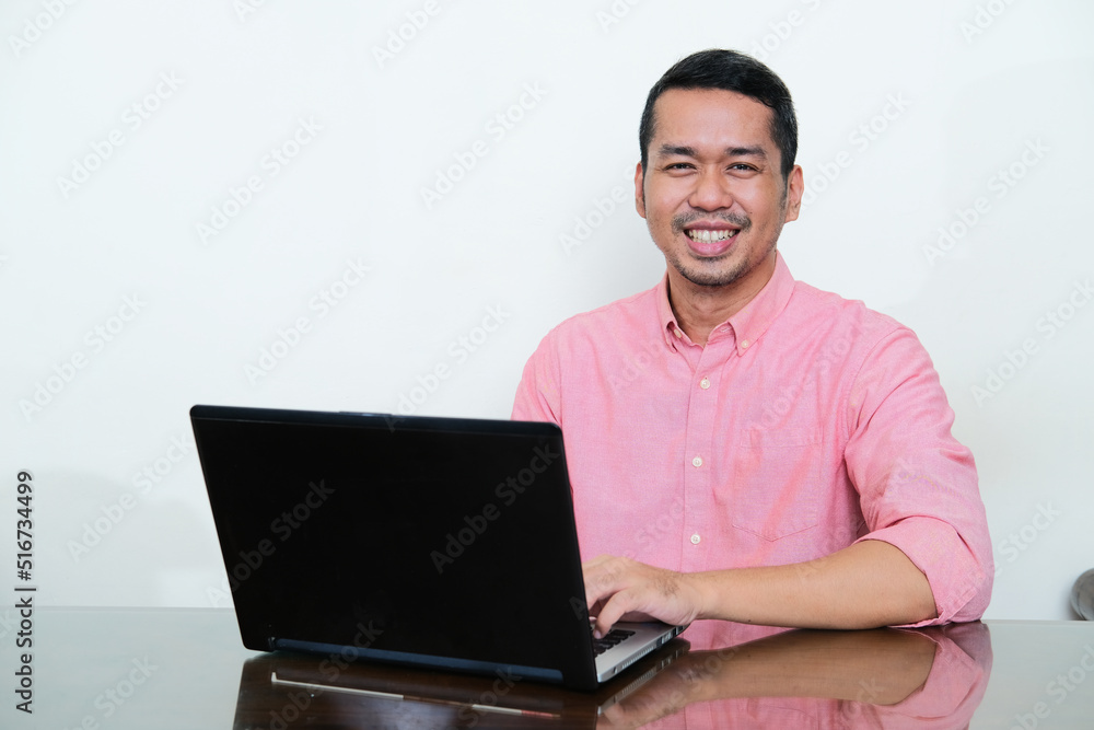 Adult Asian man sitting in front of his laptop showing happy expression Adult Asian man sitting in front of his laptop showing happy expression