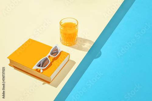 Creative summer scene with orange book, stylish white sunglasses and crystal glass on sunny blue and sandy background. Leisure by the pool idea.