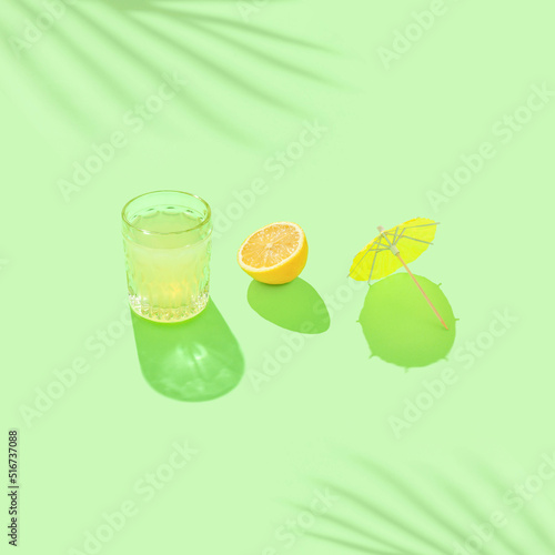 Minimal summer scene with beverage glass, halved lemon, yellow paper parasol and soft palm shadow on sunny green background.