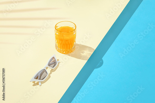 Minimal tropical composition with stylish white sunglasses and orange drink on duo tone sandy and blue background. Creative copy space with palm leaf shadow. Pool party concept.