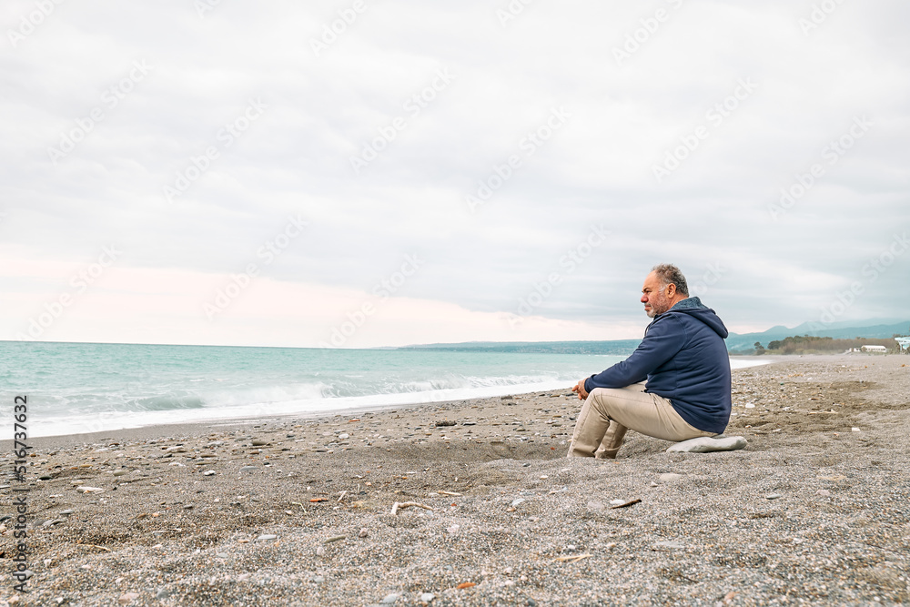 Side view of thoughtful bearded mature man sitting on pebble winter beach and contemplating blue sea. Concept of leisure activities,tourism, lifestyle e nature. Side view of thoughtful bearded mature man sitting on pebble winter beach and contemplating blue sea. Concept of leisure activities,tourism, lifestyle e nature.