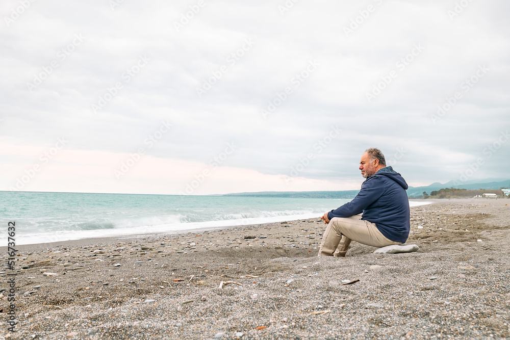 Side view of thoughtful bearded mature man sitting on pebble winter beach and contemplating blue sea. Concept of leisure activities,tourism, lifestyle e nature.