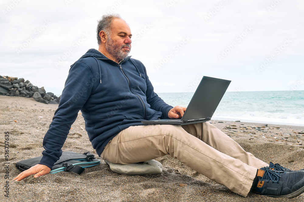 Bearded middle-aged man with laptop computer working outdoors while sitting on winter beach in front of the sea. Modern lifestyle, connection, blogging, business, freelance work.