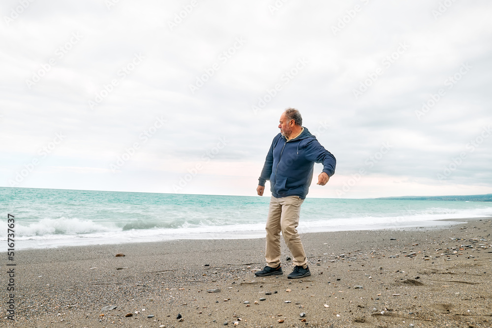 Happy middle-aged bearded man throws pebbles into the sea, walking along deserted winter beach in cloudy day. Concept of leisure activities, wellness, freedom, tourism, healthy lifestyle and nature.