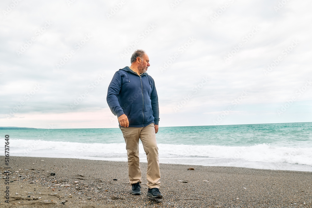 Happy middle-aged bearded man walking along deserted winter beach in cloudy day. Concept of leisure activities, wellness, freedom, tourism, healthy lifestyle and nature.