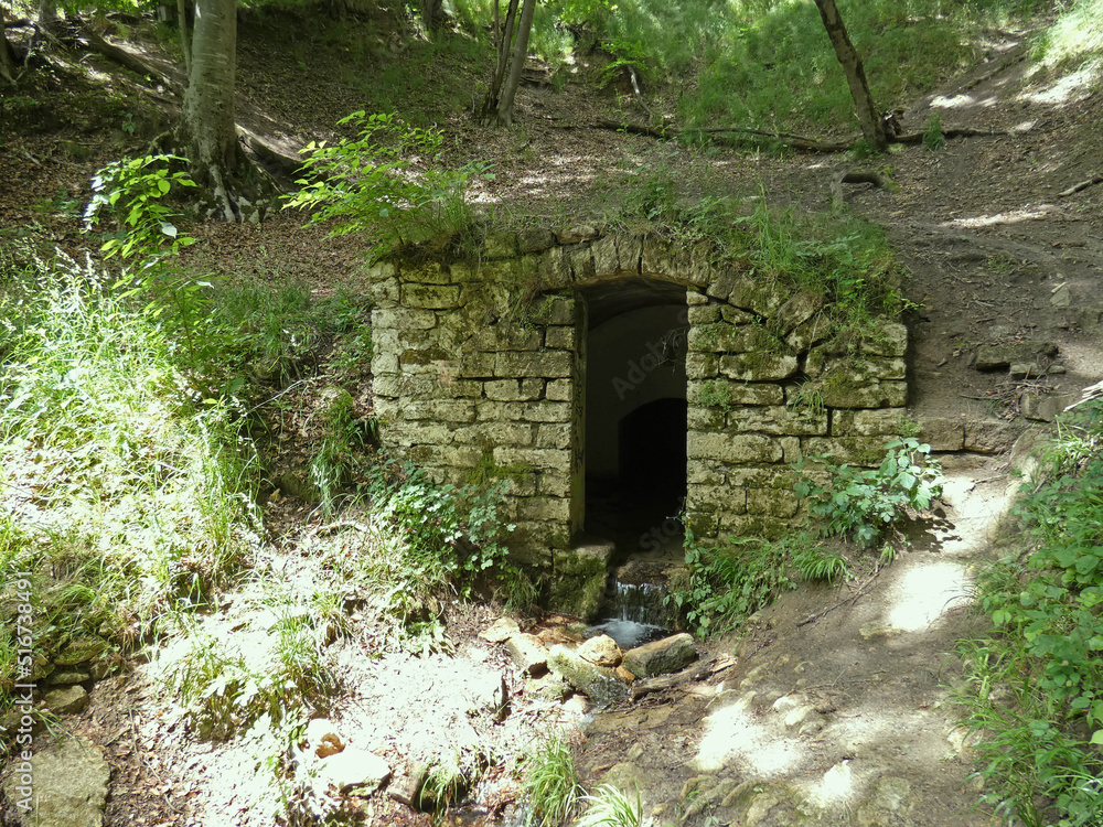 Mysterious stone door in the middle of the forest. Dark portal to the hill among old rocks. Broken trees and moss around. Small creek on the ground. Sunny day. High quality photo. Mysterious stone door in the middle of the forest. Dark portal to the hill among old rocks. Broken trees and moss around. Small creek on the ground. Sunny day. High quality photo.