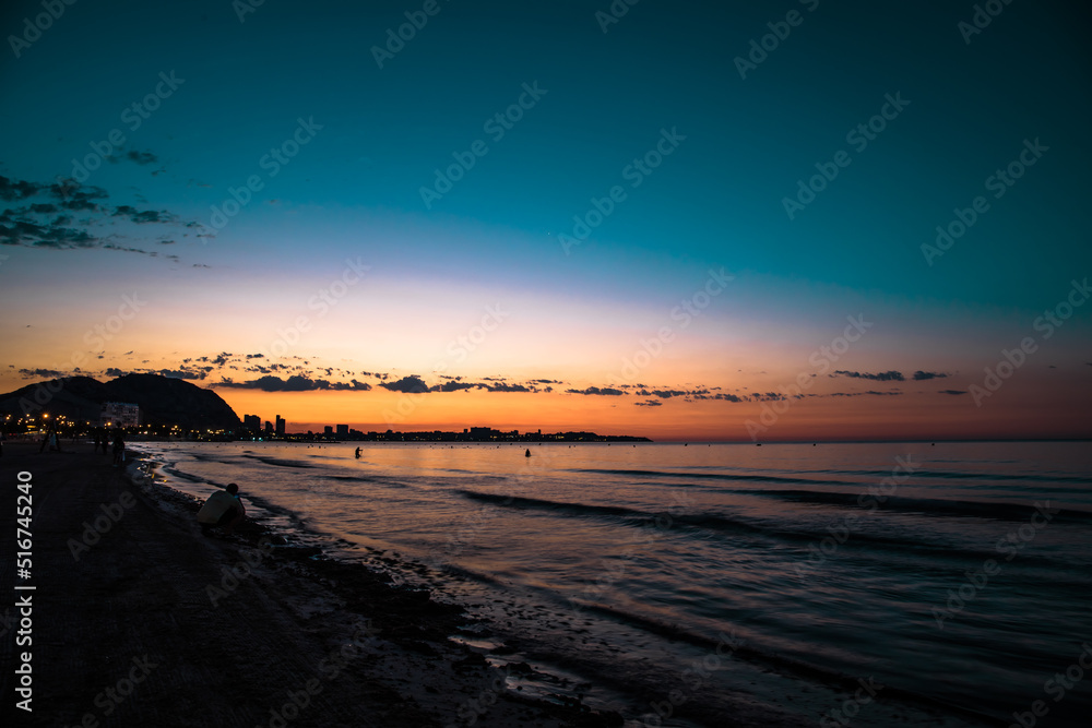 The sunrise seen from the postiguet beach in Alicante, with buildings and people against the light.