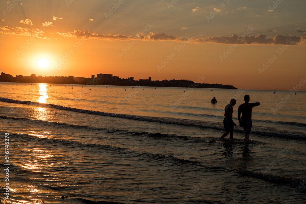 The sunrise seen from the postiguet beach in Alicante, with buildings and people against the light.