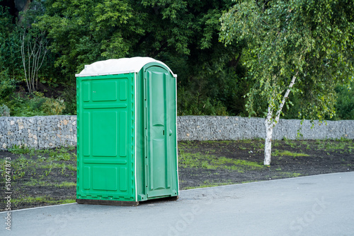 An outdoor bio toilet stands on the sidewalk in a city park, a green plastic toilet, a cabin for wc.