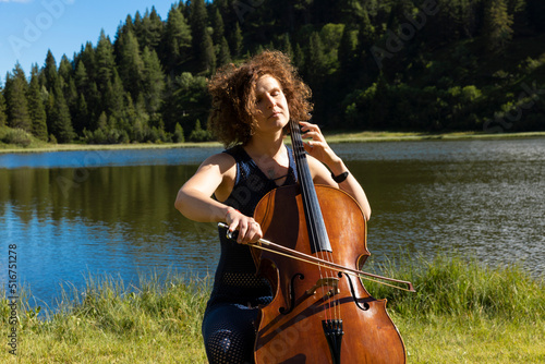 Billede på lærred Beautiful woman plays the cello in the mountains in the middle of a meadow near a lake