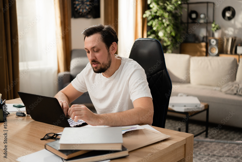 The brunette is working on laptop on new project. It's sunny day outside and the guy is in good mood. There are books and stationery on the table. The brunette is working on laptop on new project. It's sunny day outside and the guy is in good mood. There are books and stationery on the table.