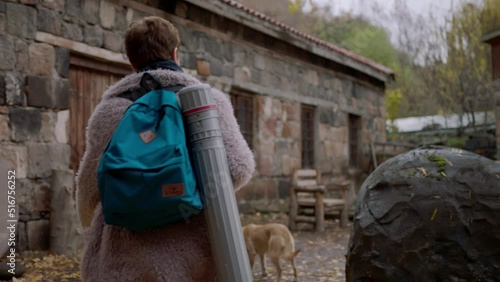 Woman's back view of walking and entering the house. Young woman designer with backpack and paper container. Dog near the house