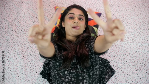 A young Indian college student making funny faces while lying down - facial expressions  funny faces. A pretty Indian female doing a thumbs-up and a victory gesture with both hands while looking at...