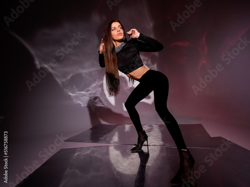 Front view of mysterious woman in black suit with crop top decorated by diamonds around neck and pants, looking away while standing on glass floor in studio with spotlights