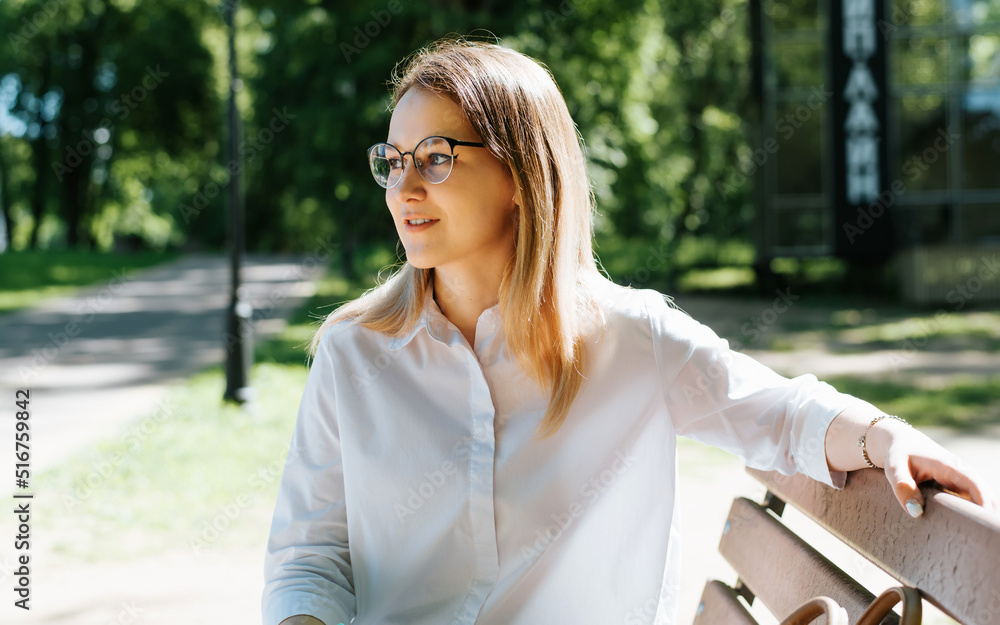 Pretty smiling young woman wearing eyeglasses and white shirt relaxing in park, looking away. Positive female student sitting alone on bench on sunny day outdoors Pretty smiling young woman wearing eyeglasses and white shirt relaxing in park, looking away. Positive female student sitting alone on bench on sunny day outdoors