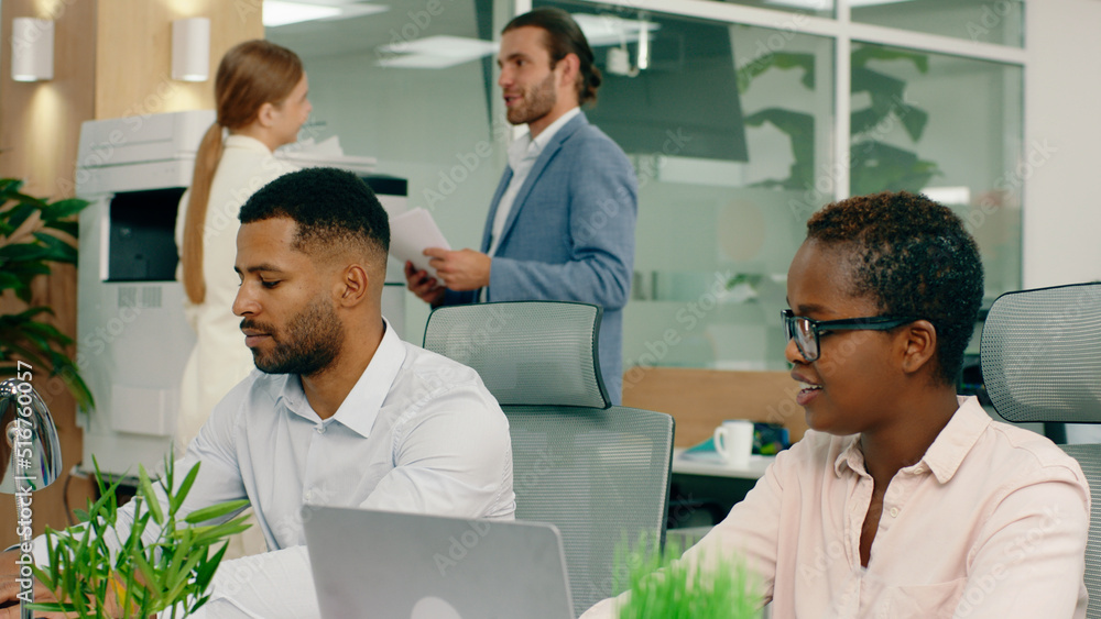 Obraz premium A black woman with short hair and glasses is talking to her colleague that s mixed race with a beard in a large lit office.