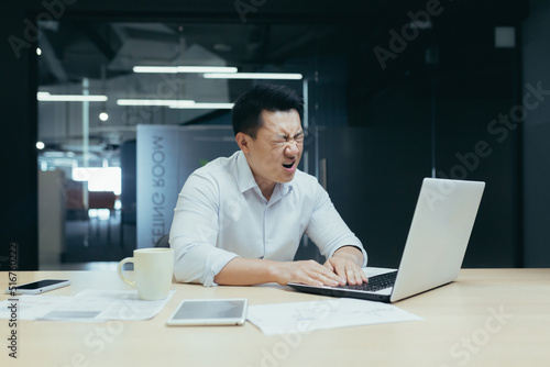 Businessman desperation, asian man working in office, looking at laptop screen, screaming in grief, got bad news