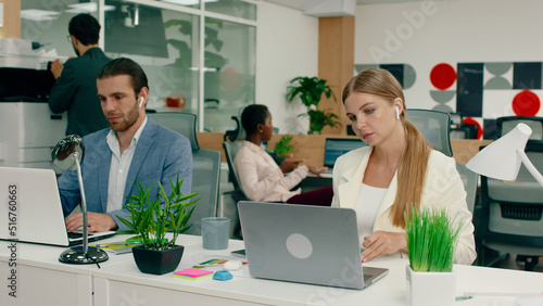 A beautiful blonde woman with her hair tied in a low ponytail, wearing a white blazer is doing work on her laptop and beside her is a handsome man in a blue suit also doing work on his laptop while