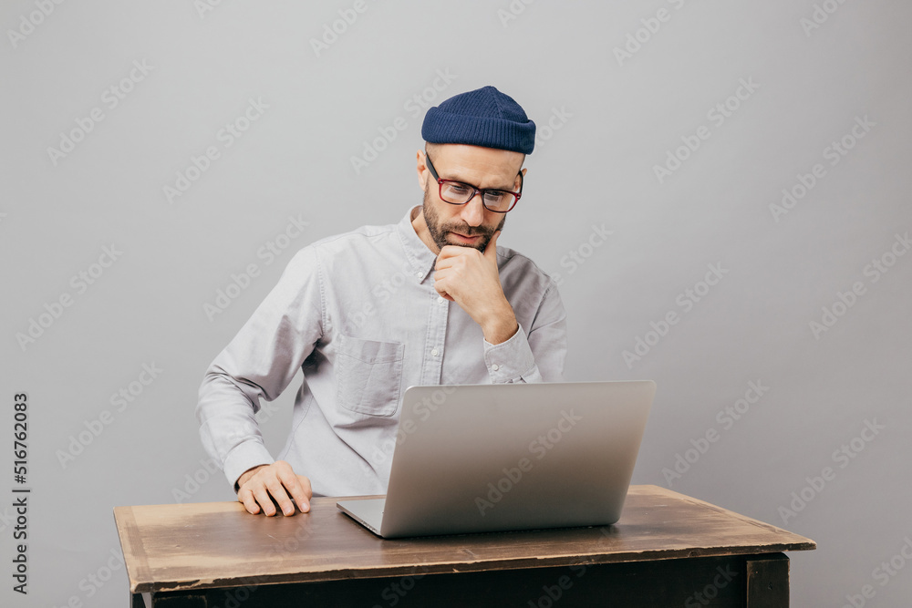 Male freelancer reads news in internet, looks attentively at laptop computer, watches video, sits at old table, wears white shirt and headgear, isolated over grey wall. Businessman ponders on startup Male freelancer reads news in internet, looks attentively at laptop computer, watches video, sits at old table, wears white shirt and headgear, isolated over grey wall. Businessman ponders on startup