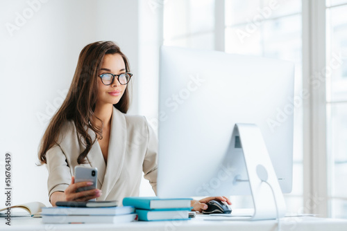 Pretty female entrepreneur with long dark hair, works on computer, develops strategy of advertising campaign, poses in modern coworking office, uses cellphone for chatting online, wears glasses