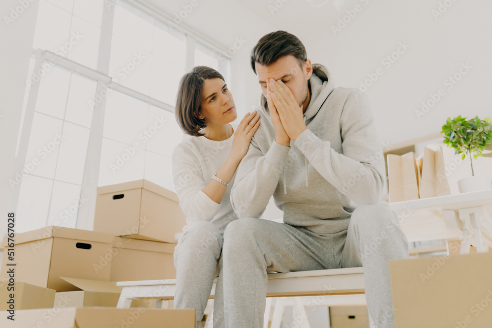 Photo of dissatisfied husband and wife forced to sell apartment because of financial problems, woman soothes husband, pose on cardboard boxes, pose in living room. Moving as kind of stress for family Photo of dissatisfied husband and wife forced to sell apartment because of financial problems, woman soothes husband, pose on cardboard boxes, pose in living room. Moving as kind of stress for family