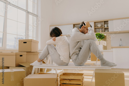 Photo of stressful couple get divorse, leave house, start living seperately, sit backs to each other, pose in kitchen, cardboard boxes with belongings around, focused down with sad expressions photo