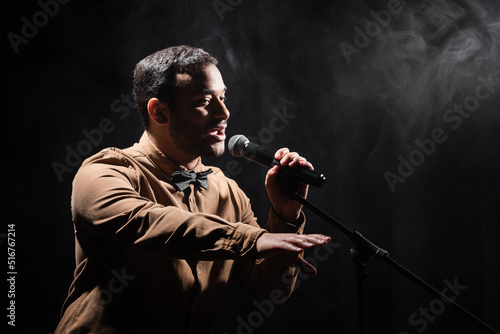 indian comedian performing stand up comedy into microphone and looking away on black with smoke.