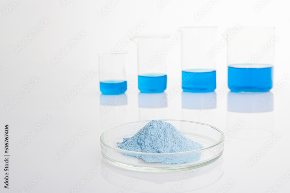 Blue biological powder in a petri dish in the foreground. In the background are laboratory flasks with blue liquid. Blue biological powder in a petri dish in the foreground. In the background are laboratory flasks with blue liquid.