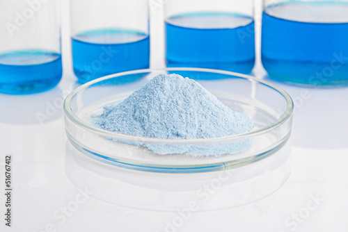 Blue biological powder in a petri dish in the foreground. In the background are laboratory flasks with blue liquid.