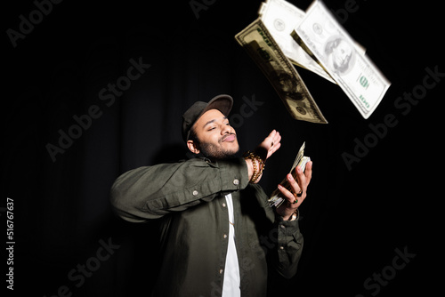 rich indian hip hop performer in cap throwing dollar banknotes on black.