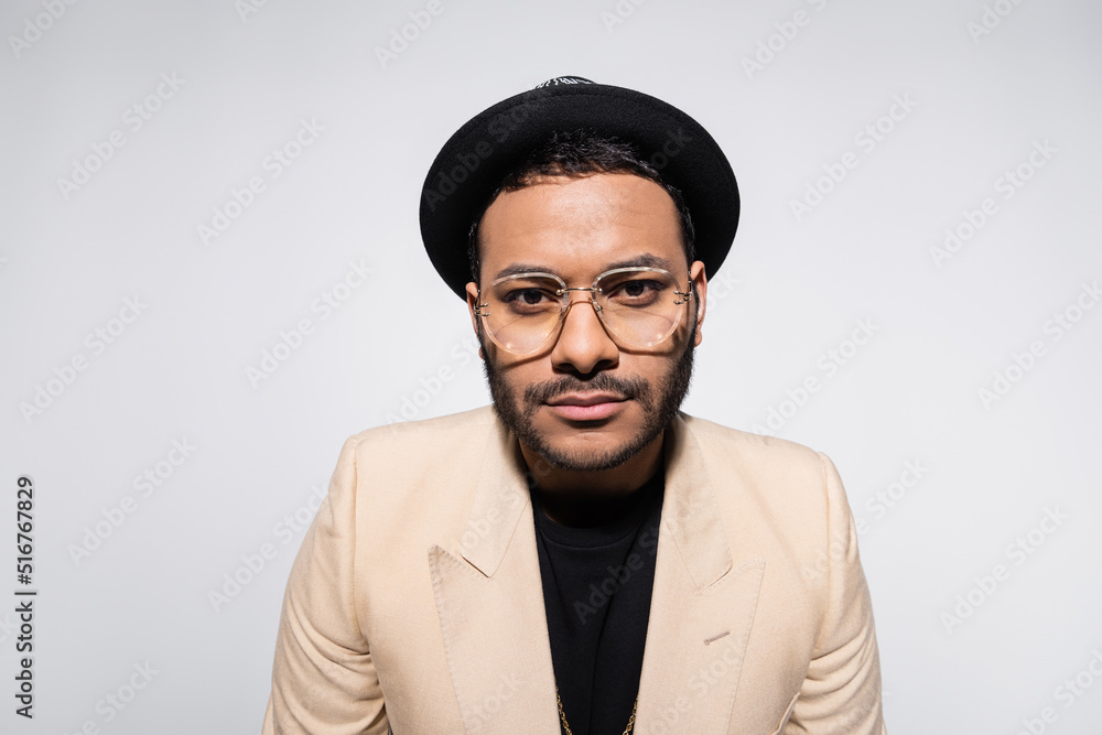 Fototapeta premium stylish indian hip hop performer in fedora hat and eyeglasses looking at camera isolated on grey.