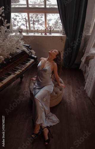 teen leisure. young brunette glamour girl in silk blue dress is sitting sensual near black grand piano with flowers and looking up on vintage room background with window. lifestyle concept, free space