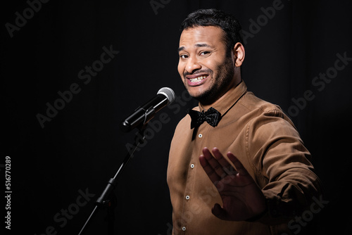 displeased indian stand up comedian showing stop gesture while telling jokes into microphone isolated on black.