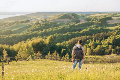 Tourist with backpack standing on top of hill in grass field and enjoying beautiful landscape view. Rear view of teenage boy hiker resting in nature. Active lifestyle. Concept of local travel © Lyubov