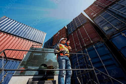 Asian man engineer Standing on a container forkift or Reach Stacker, using a communication radio Walkie-Talkie, with cotainers backgroud, to engineer and transport industry concept.