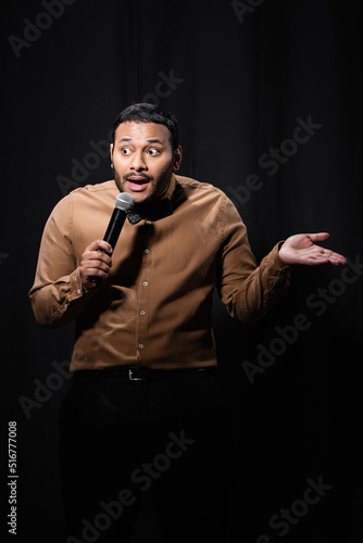 emotional indian comedian in shirt and bow tie holding microphone and gesturing during monologue on black.