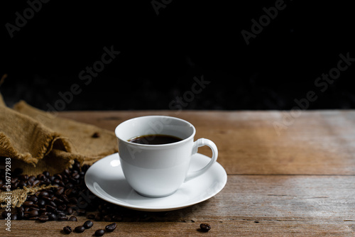Hot coffee in a white coffee cup and many coffee beans placed around and on a wooden table in a warm, light atmosphere, on dark background, with copy space.