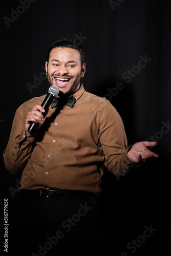 joyful indian comedian in shirt and bow tie holding microphone during monologue on black.