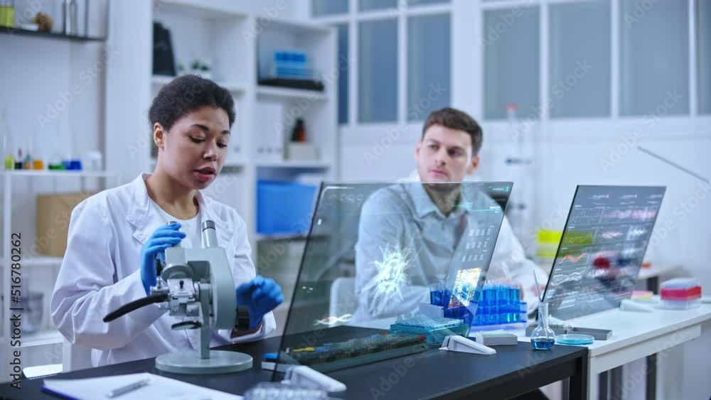 Female scientist examining cell under microscope, male assistant writing results Female scientist examining cell under microscope, male assistant writing results