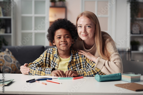 Portrait of young foster mother looking at camera with her adoptive son while they sitting at table at home