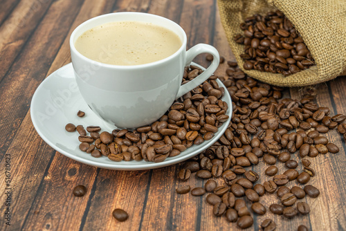 A cup of creamy coffee with roasted coffee beans next to it, wooden background